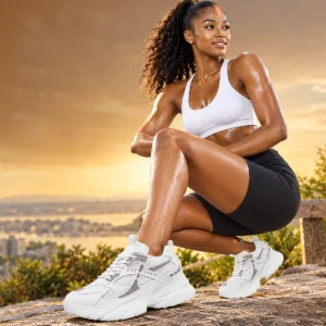 A woman wearing a white sports bra and black biker shorts squats outdoors while wearing Acuvick mesh shoes during a sunset workout.